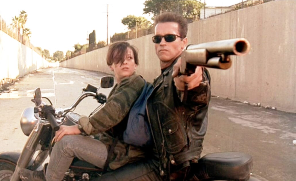 Movie still: a man in sunglasses and a leather jacket rides a motorcycle down a concrete storm channel with a boy behind him, aiming a shotgun ahead