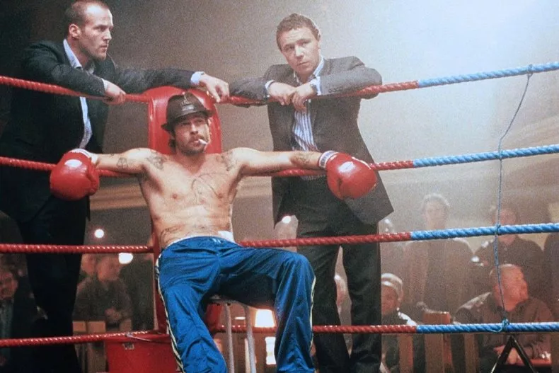 Still from the film Snatch: a bare-chested boxer in a flat cap slumps on his stool in the corner of the ring while two men in suits lean on the ropes behind him