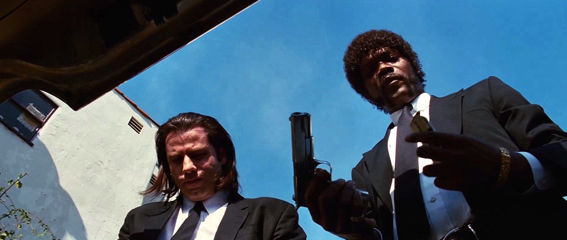 Two men in black suits look down into an open car trunk, one holding a pistol, filmed from inside the trunk