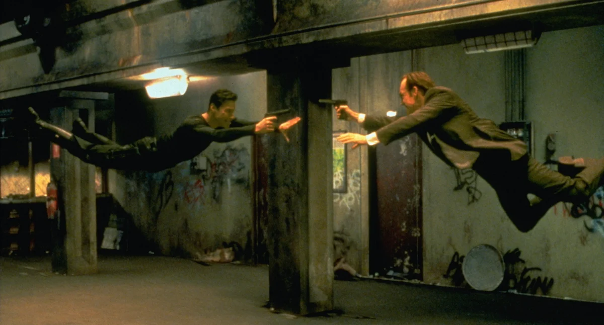 Still from The Matrix — two characters diving with guns drawn past a concrete pillar in a car park