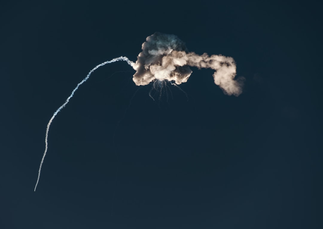 A rocket trail curving upward then exploding into a cloud of smoke against a dark blue sky